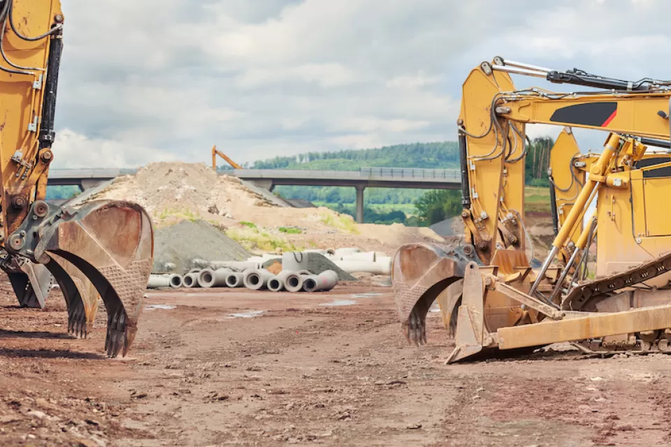 Three yellow excavators at a road construction site with elevated roadway in background.
